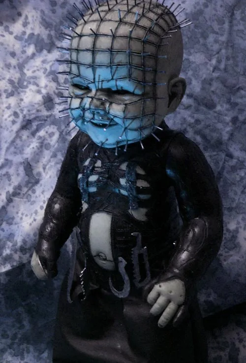A small, doll-like figure resembling the horror character Pinhead, with a blue-painted face and head covered in a grid of…