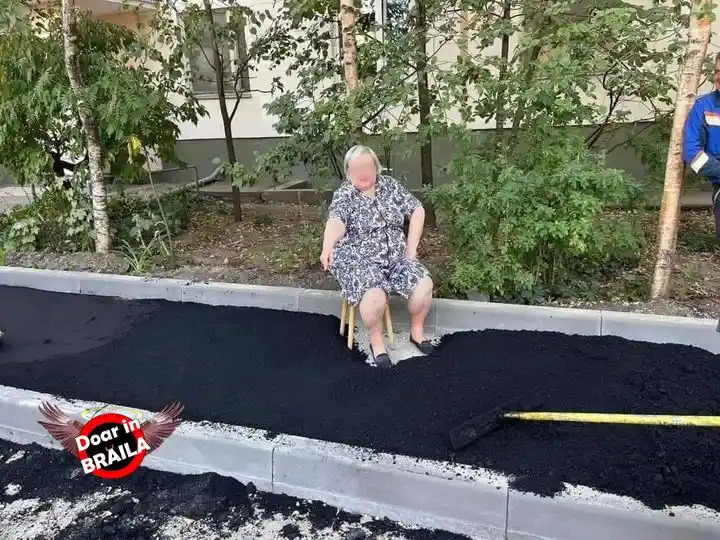 An older woman sits on a small wooden stool in the middle of a freshly paved asphalt road, surrounded by greenery and a…