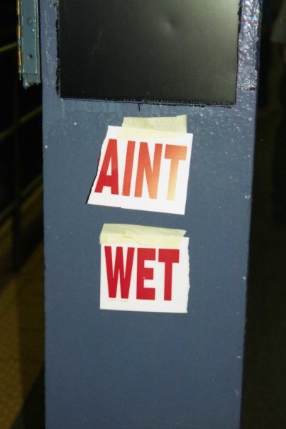 Two red-and-white stickers are taped vertically on a blue metal pole, reading 'AINT' above 'WET'. The stickers are slightly…