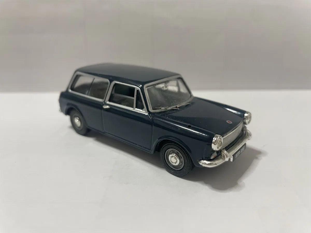 A dark blue die-cast model of a vintage station wagon car, likely a Mini, is photographed on a plain white surface against a…