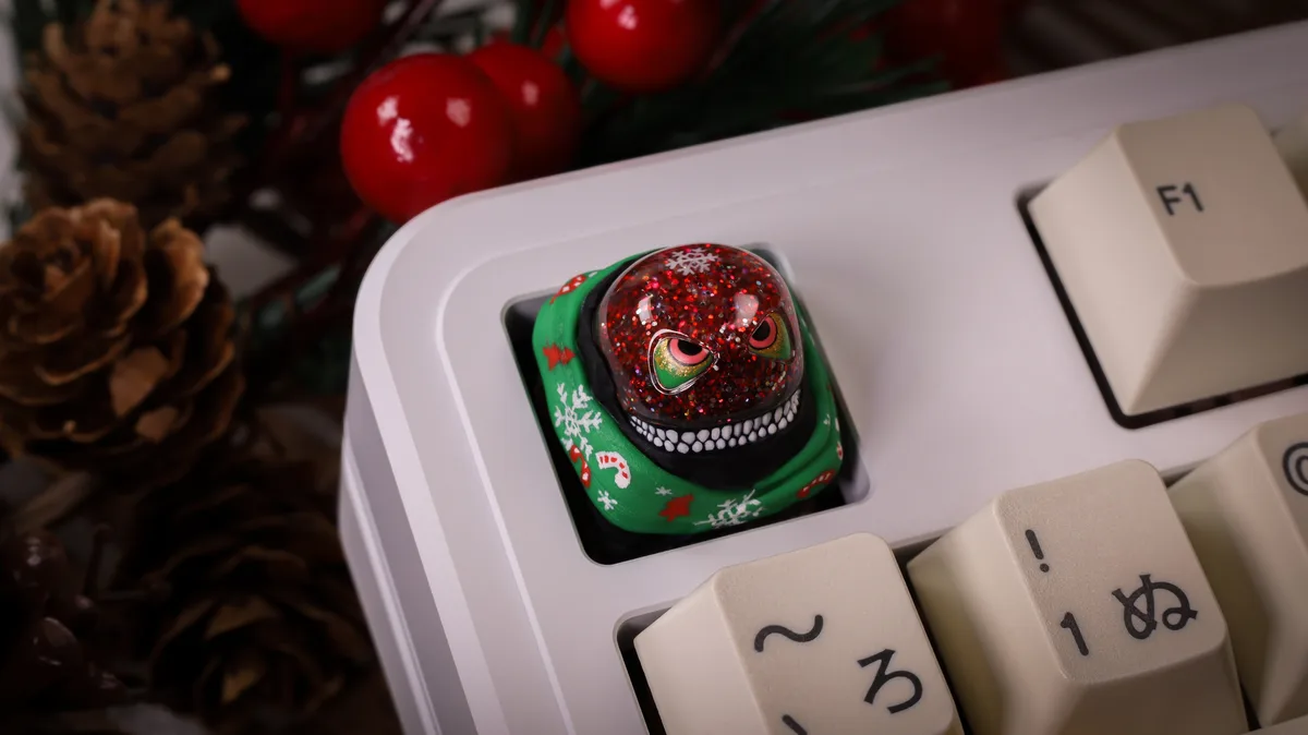 a close-up photo shows a custom keyboard keycap designed as a festive, menacing face with red glitter, green…