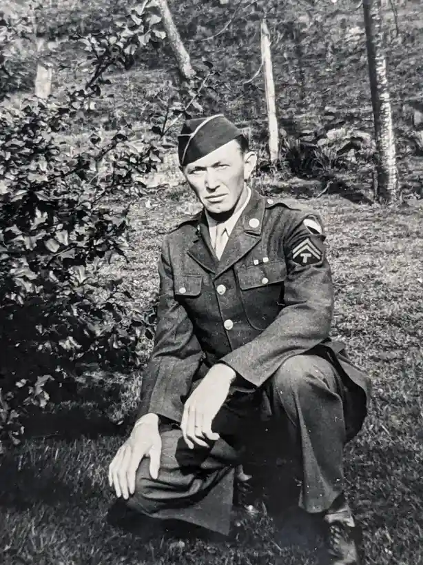 A black-and-white photograph shows a man in a U.S. Army uniform from the mid-20th century, crouching outdoors in a wooded…
