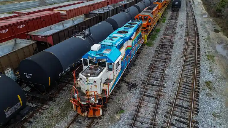 An aerial view shows a blue and white locomotive labeled 'BUGX RAIL' with number 814, pulling black tanker cars on a railway…