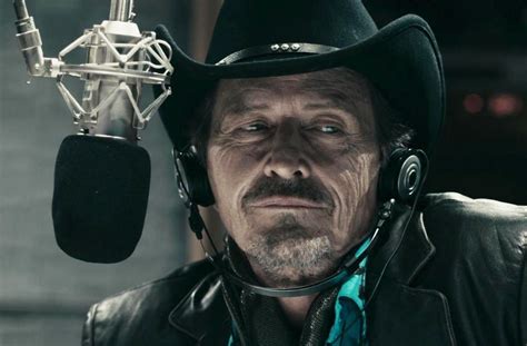A close-up of a man with a weathered face, wearing a black cowboy hat, headphones, and a black leather jacket, sitting in…