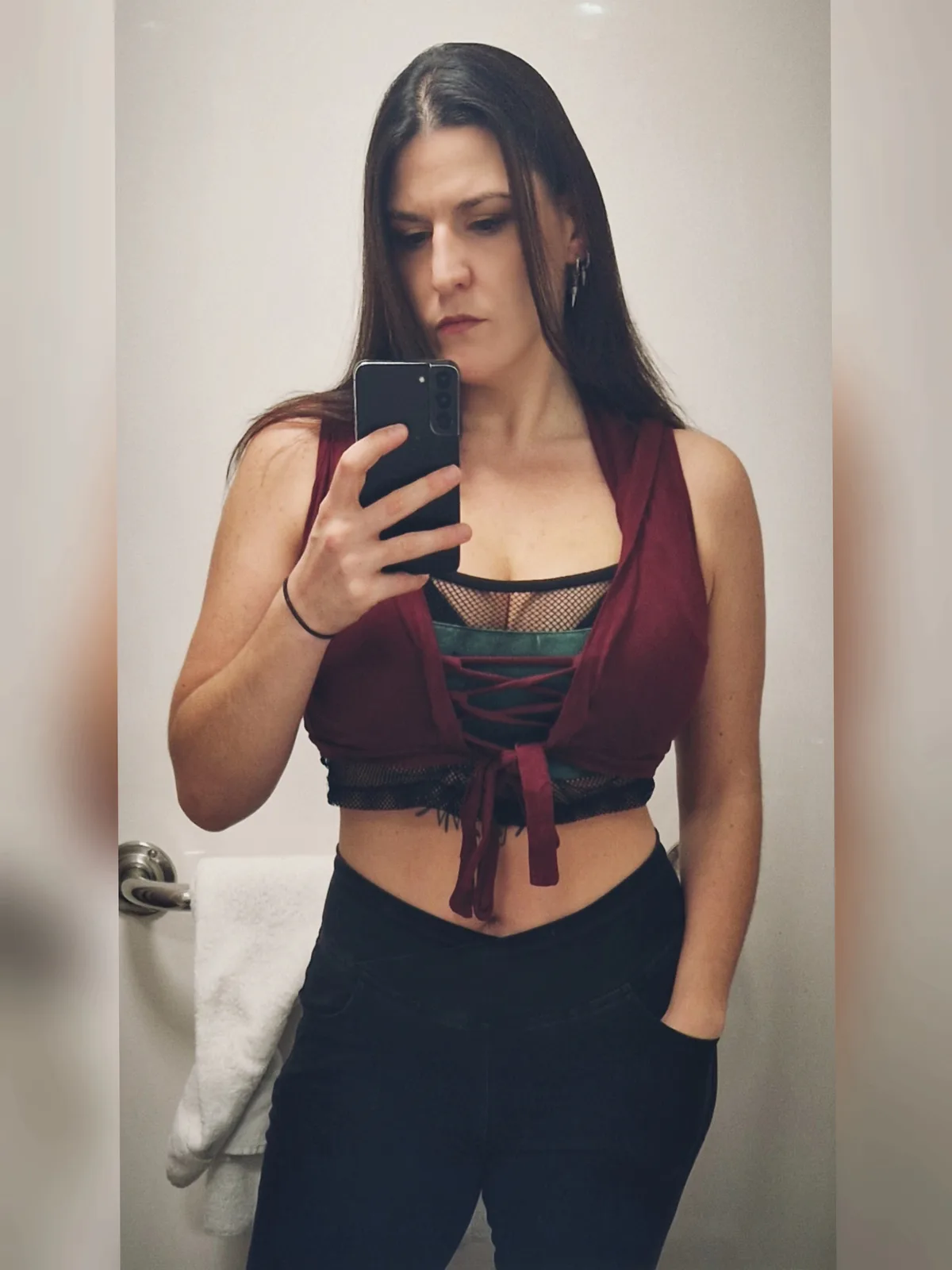 A woman with long dark hair takes a mirror selfie in a bathroom, wearing a layered outfit with a maroon crop top over a…