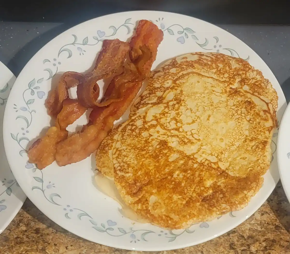 A close-up photo shows a plate with two golden-brown pancakes and a serving of crispy bacon, both resting on a white plate…