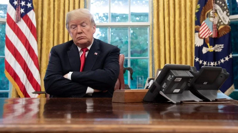 a man in a dark suit and red tie sits at a wooden desk with arms crossed, looking displeased. Behind him are yellow…