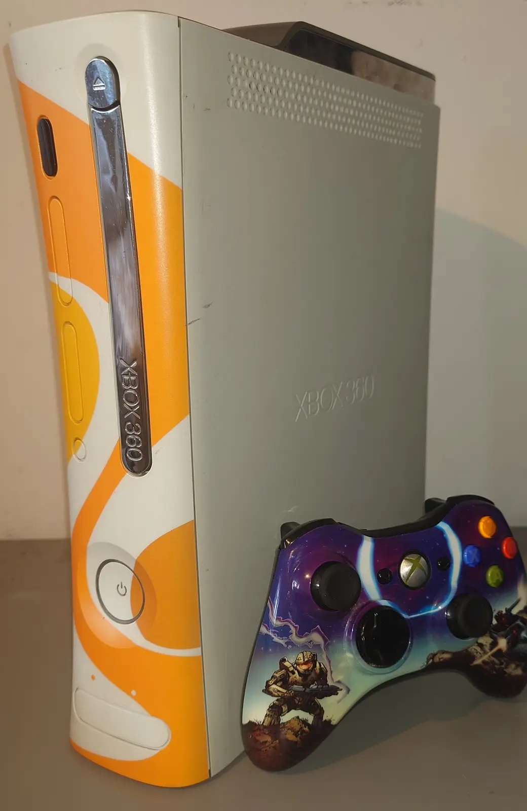 A white Xbox 360 console with orange and white swirl decals stands next to a custom controller featuring a Halo Spartan and…