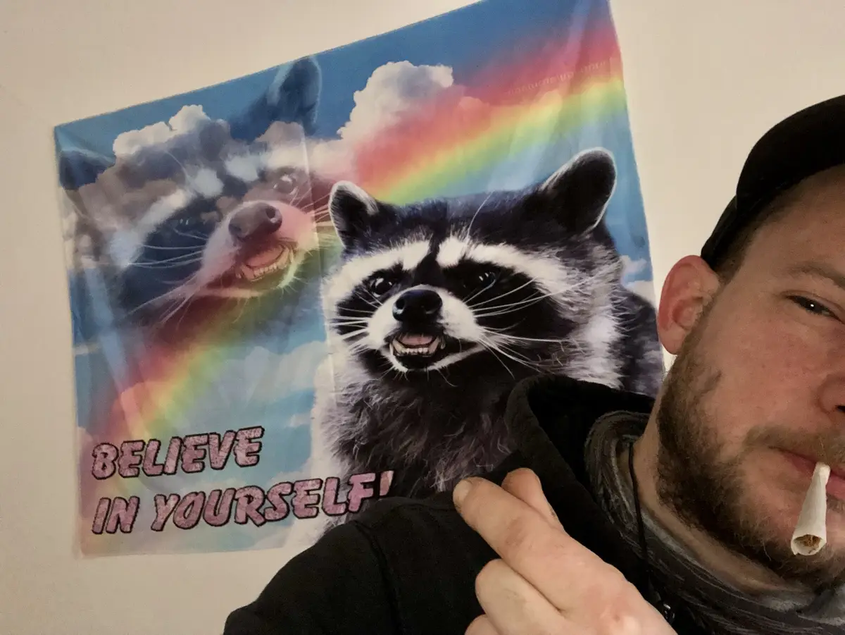 A person with a beard and a black cap is taking a selfie, holding a rolled cigarette in their mouth, next to a colorful…