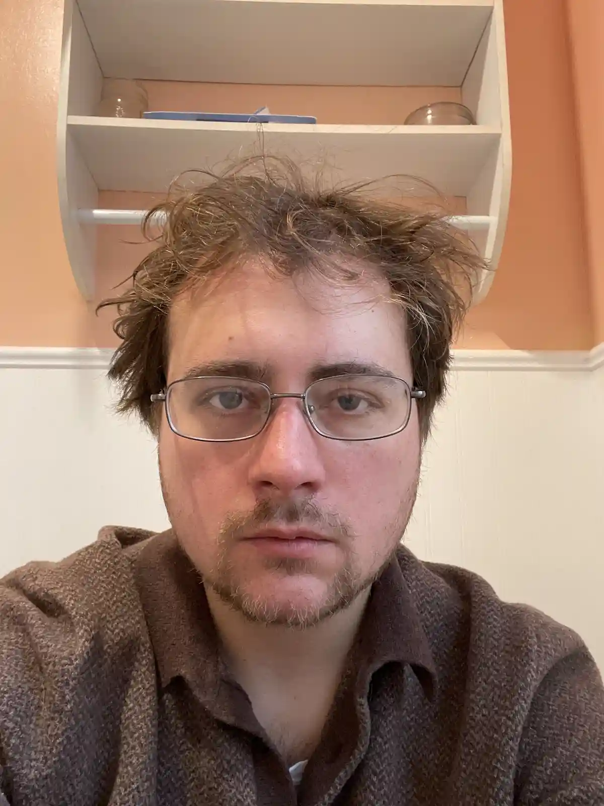 A close-up selfie of a man with messy brown hair, glasses, and a brown textured shirt, looking directly at the camera with a…
