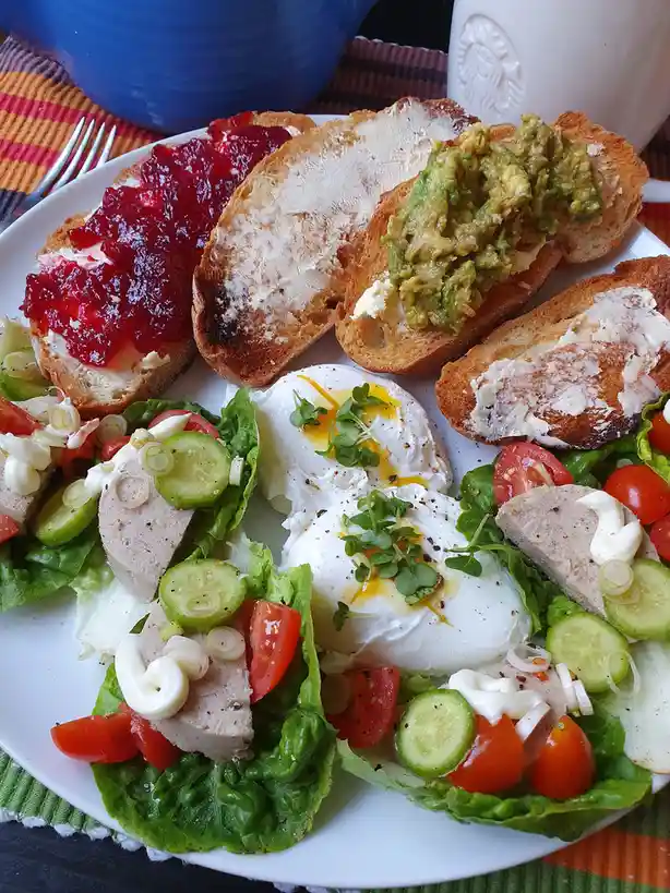 A plate of healthy breakfast food including avocado toast, jam toast, poached eggs, and a salad with chicken, tomatoes…