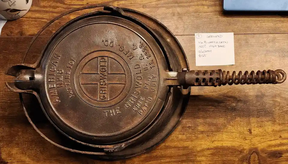 A vintage Griswold No. 8 cast iron waffle iron rests on a wooden table, its lid showing embossed branding and manufacturing…