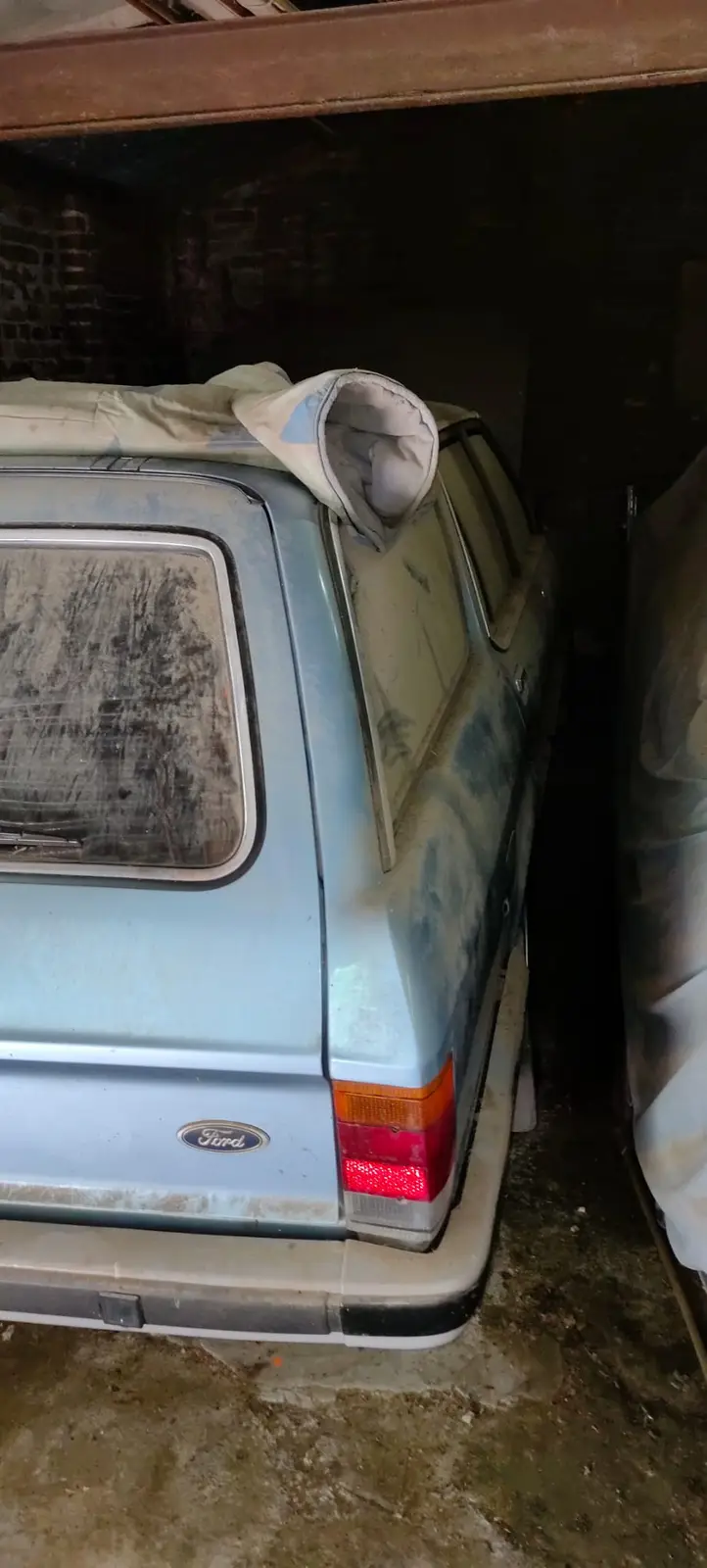 A dusty, light blue Ford station wagon is parked in a dim, cluttered garage or storage area, covered partially by a tarp…
