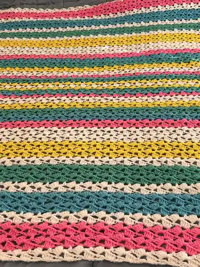 A close-up photo of a handmade, multi-colored crocheted blanket with repeating horizontal stripes in pink, teal, green…