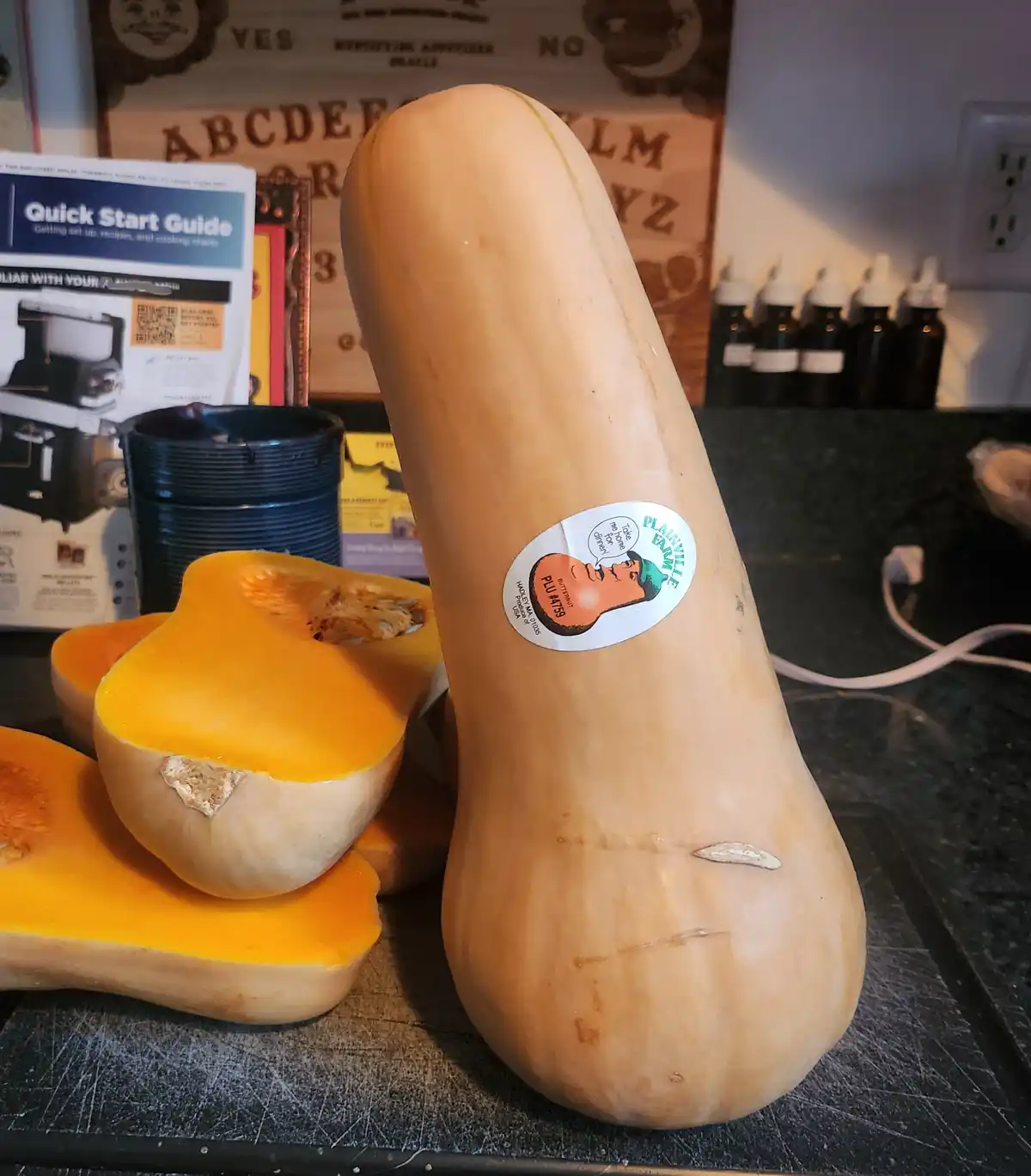 A large butternut squash sits upright on a kitchen counter next to several cut halves, its sticker featuring a cartoon face…