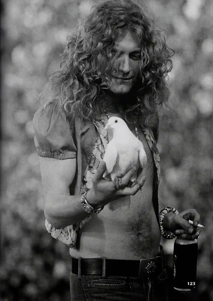 A black-and-white photo shows a man with long curly hair holding a white dove in one hand and a cigarette in the other…