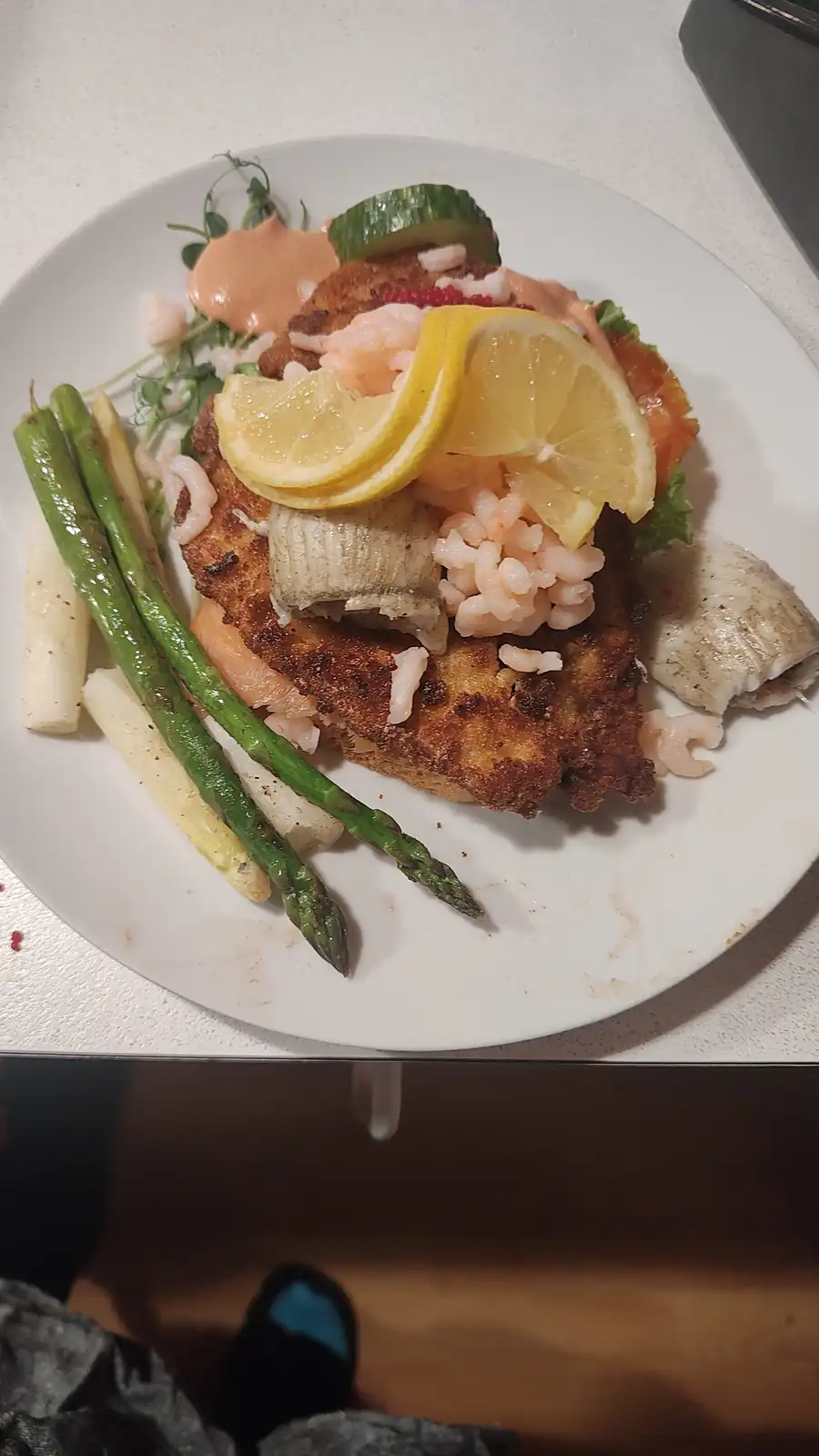 A plate of food featuring a breaded and fried fish fillet topped with shrimp, lemon slices, cucumber, and sauce, accompanied…