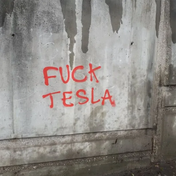 A close-up of a weathered concrete wall with visible water stains and grime, featuring the phrase 'FUCK TESLA' spray-painted…