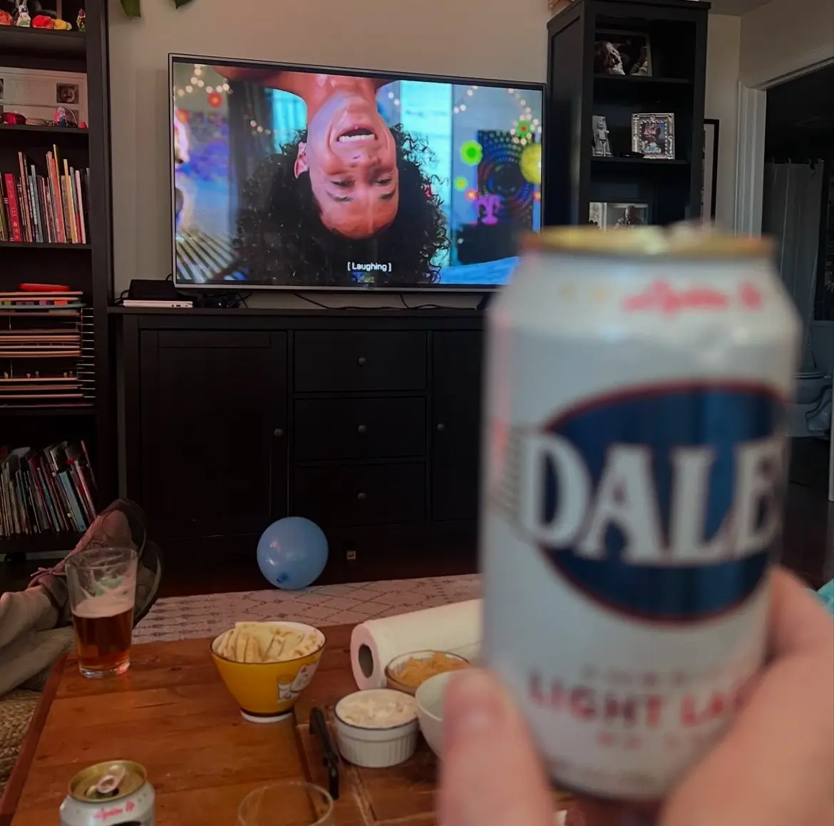 A person holds a blurry can of Dale's Light Lager in the foreground, obscuring most of the view. In the background, a TV…