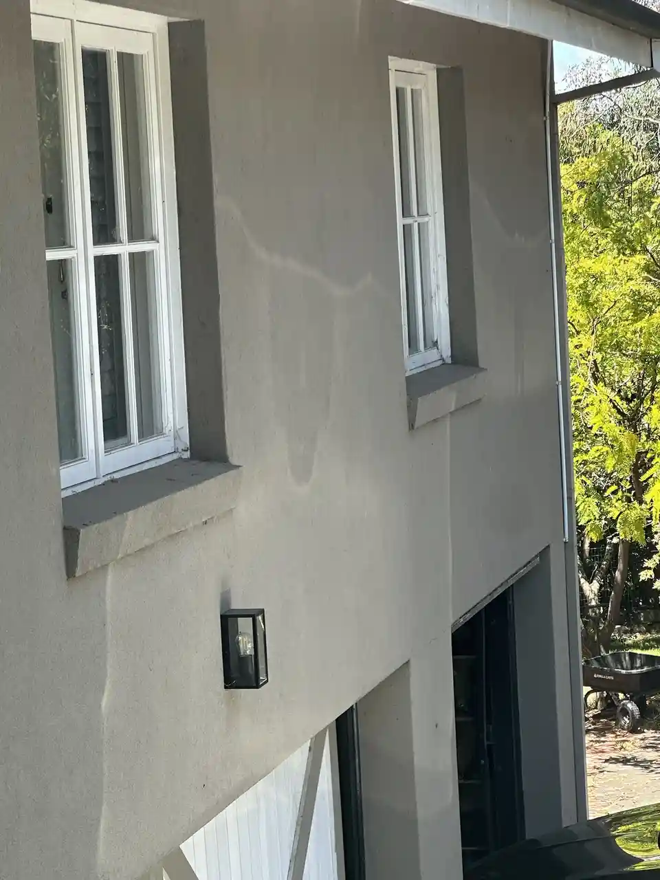A photo of a plain gray house exterior with two white-framed windows and a small black outdoor light fixture. The sun casts…