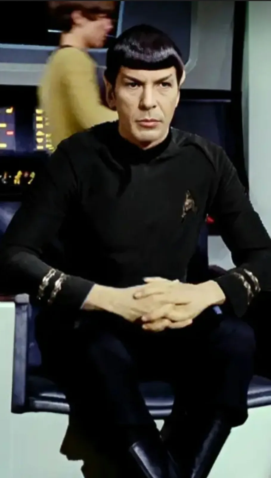 A still image of the character Spock from Star Trek, seated in a chair with a serious, unimpressed expression. He wears his…