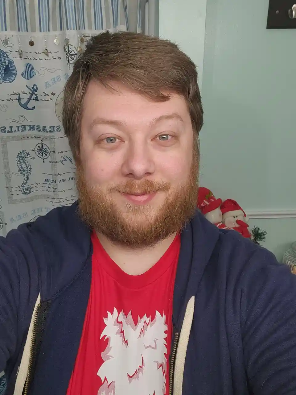 A man with a reddish beard and blue eyes smiles at the camera while wearing a red t-shirt under a navy blue zip-up hoodie…
