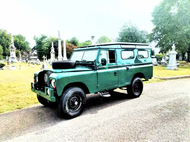 A green vintage Land Rover is parked on a gravel path inside a cemetery, surrounded by gravestones and monuments under a…