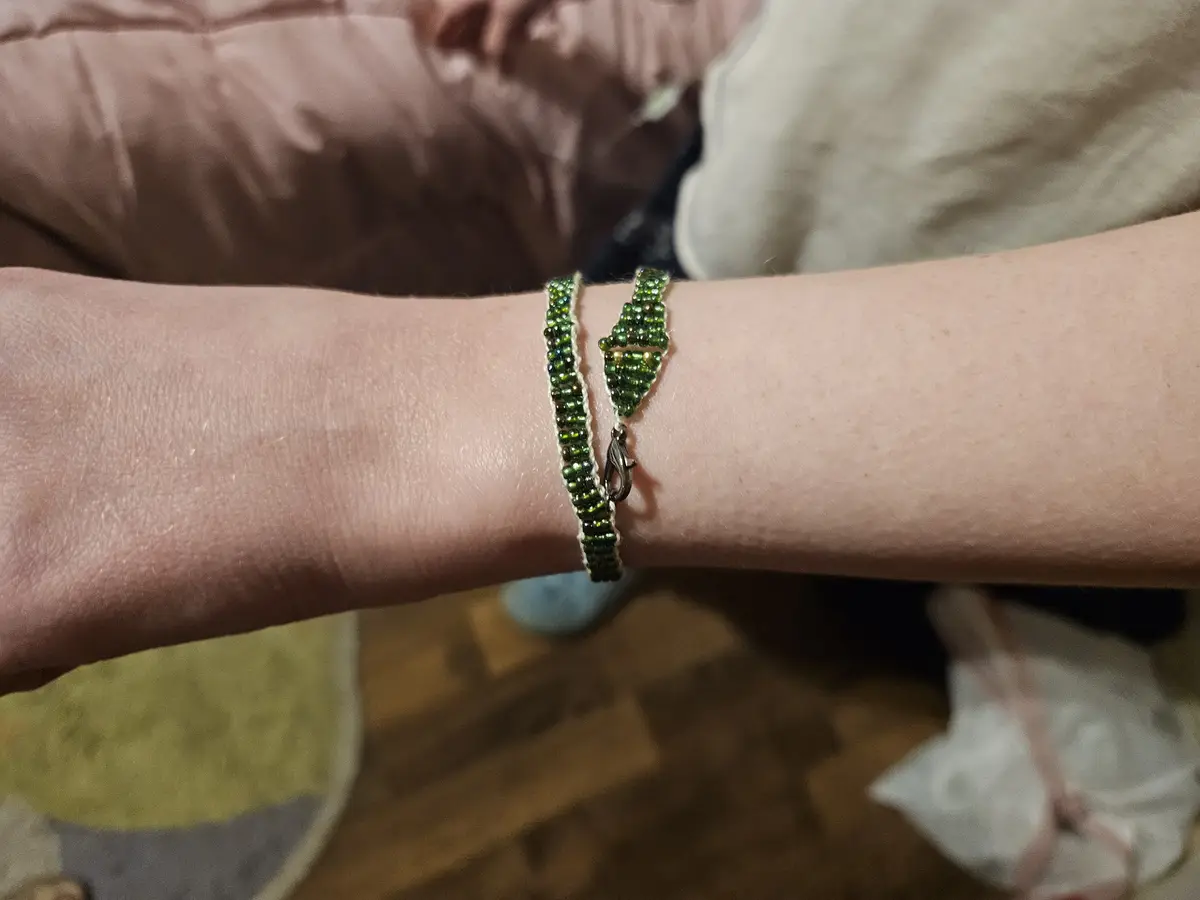 A close-up photo shows a person's wrist wearing a handmade green beaded bracelet with a silver clasp. The bracelet has a…
