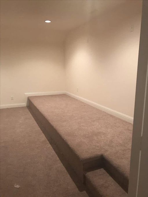 An empty room with beige carpeting features a raised platform that looks like a bed but has no mattress or bedding, with…