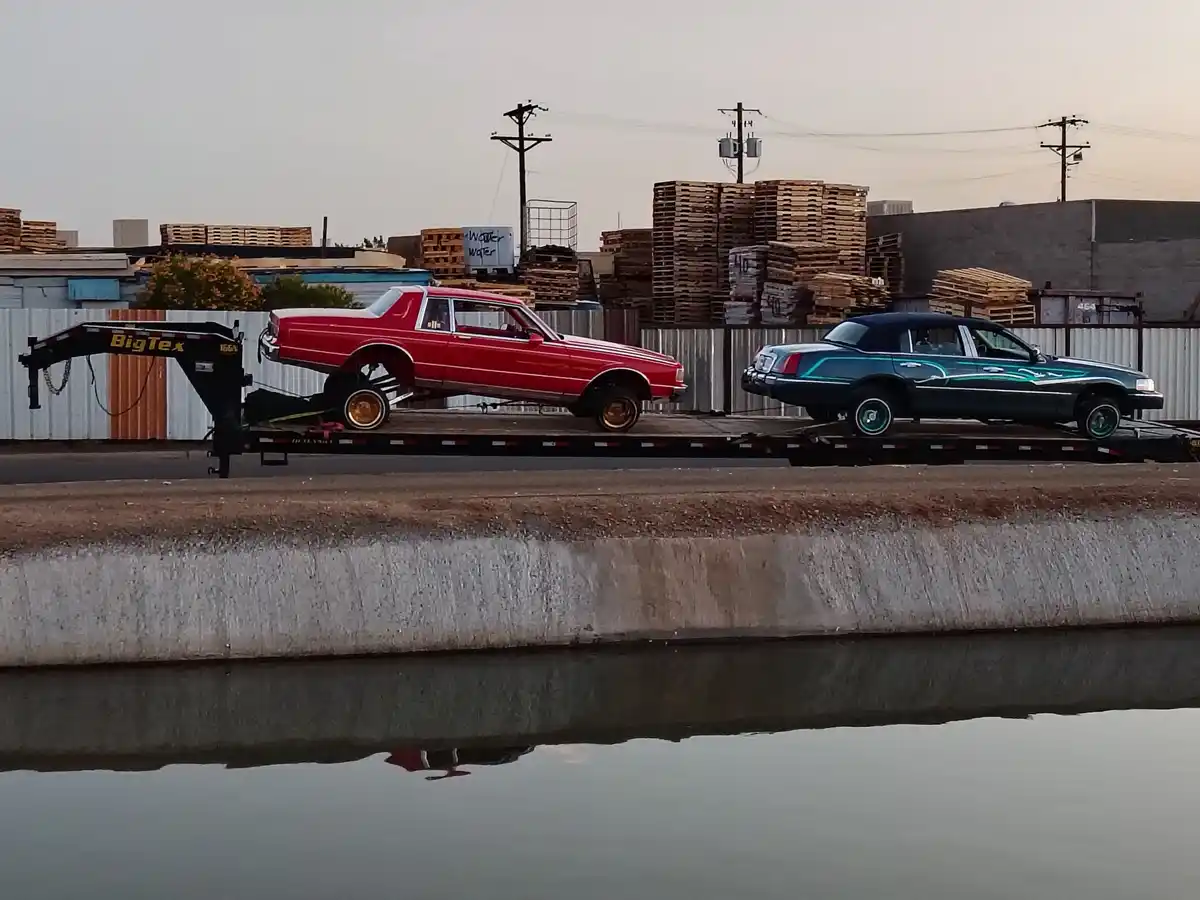 A flatbed trailer carries two cars: a red classic sedan with gold rims and a teal sedan with custom paint, both elevated on…