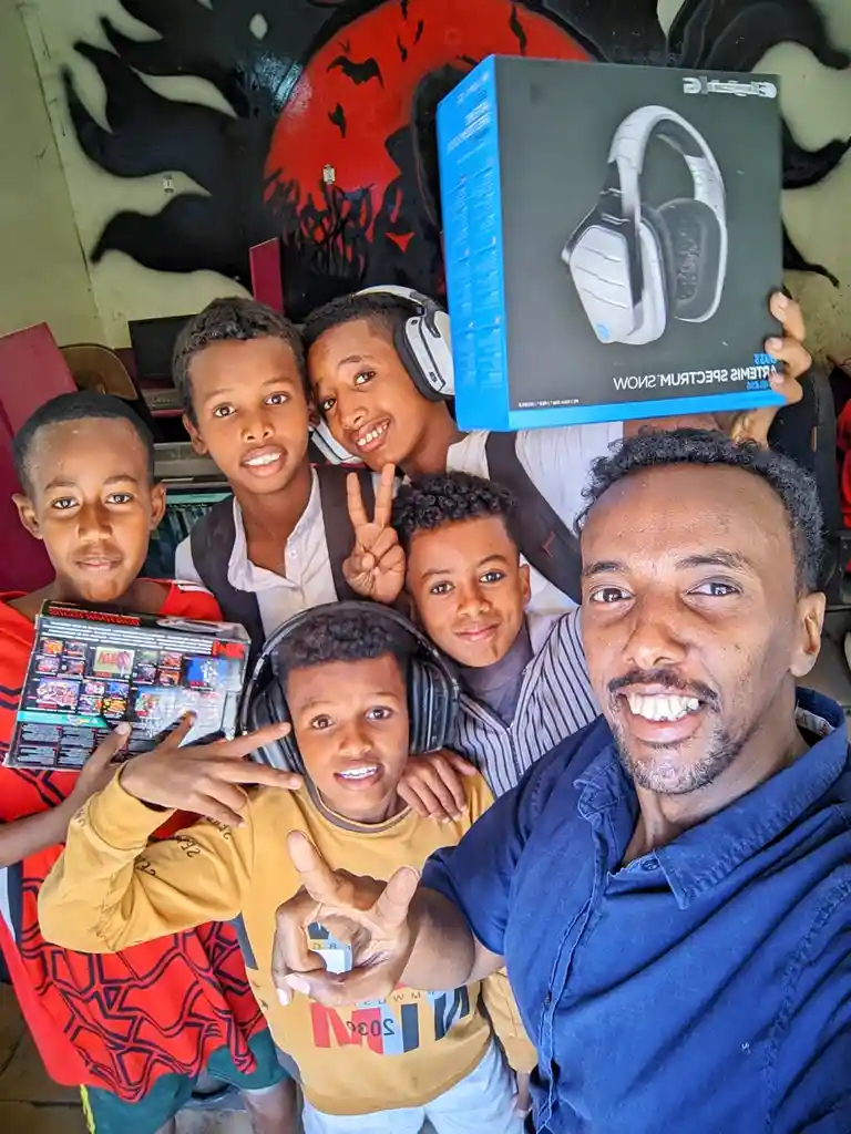 A man takes a selfie with five young boys in front of a graffiti wall. One boy holds a box of Logitech headphones, another…