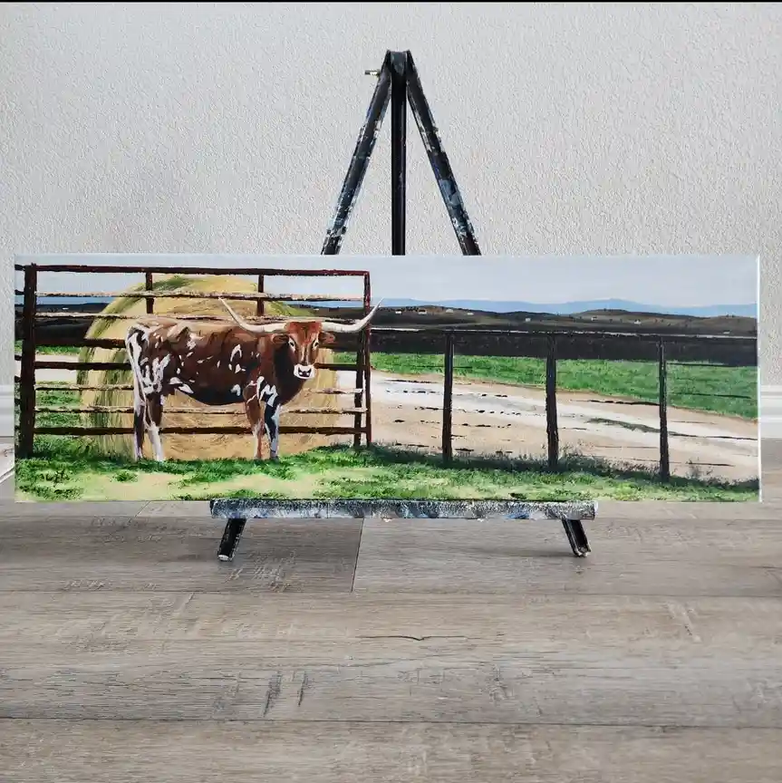 A painting of a brown and white longhorn cow standing in a fenced pasture with hay bales, displayed on an easel indoors on a…