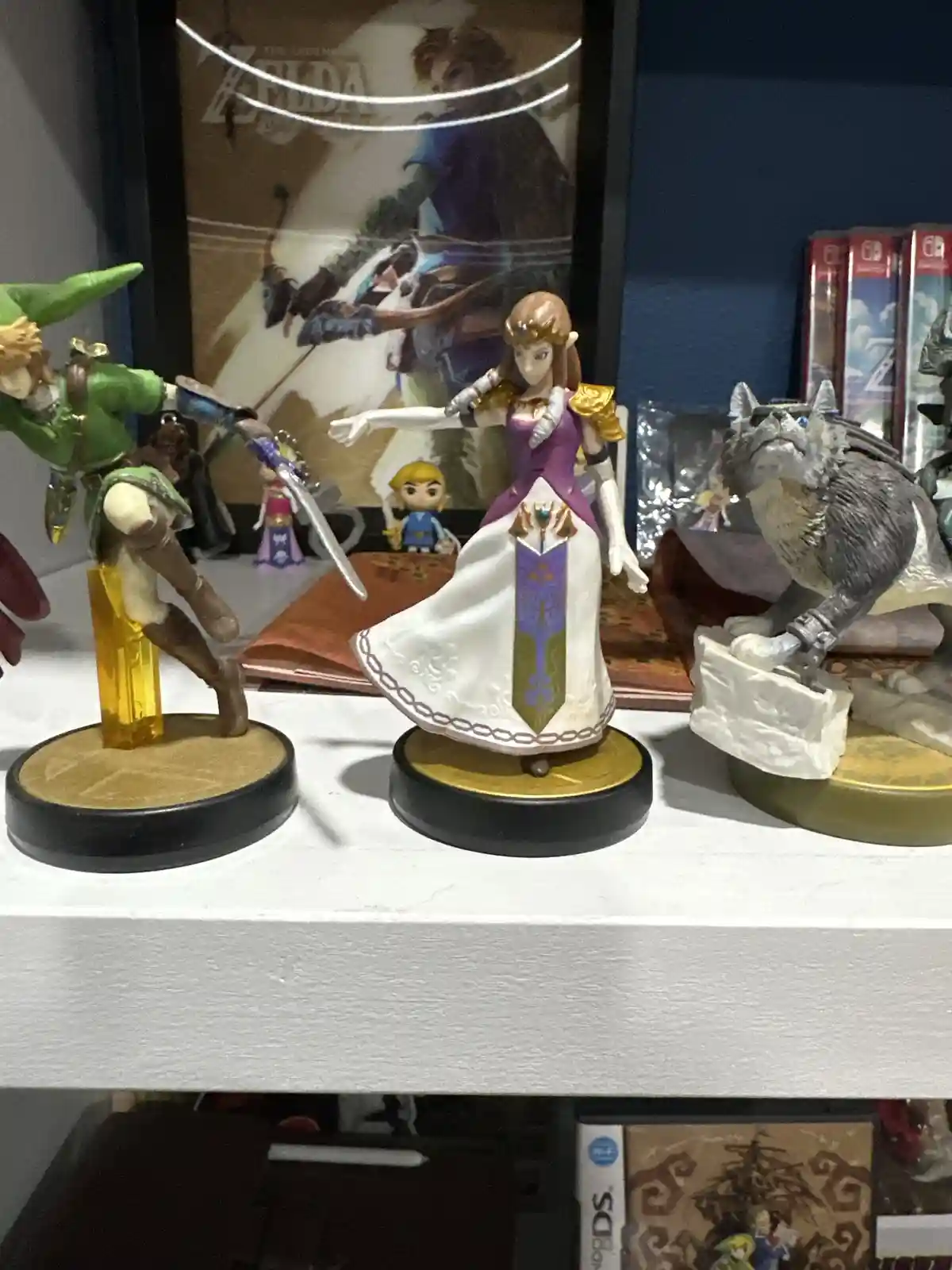 Three amiibo figures from The Legend of Zelda series are displayed on a shelf: Link in green tunic holding a sword, Zelda in…