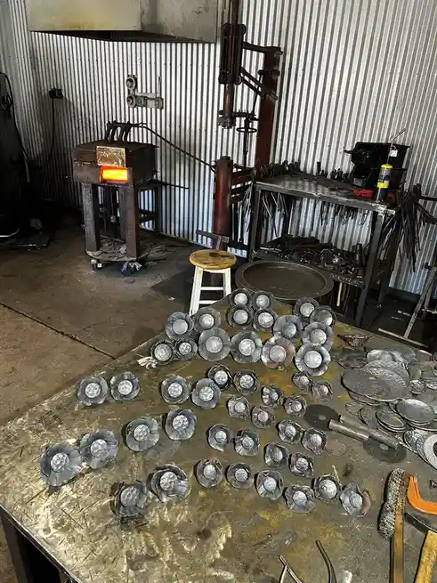 A metalworking workshop features a workbench covered with dozens of handmade, flower-shaped metal pieces, with a glowing…
