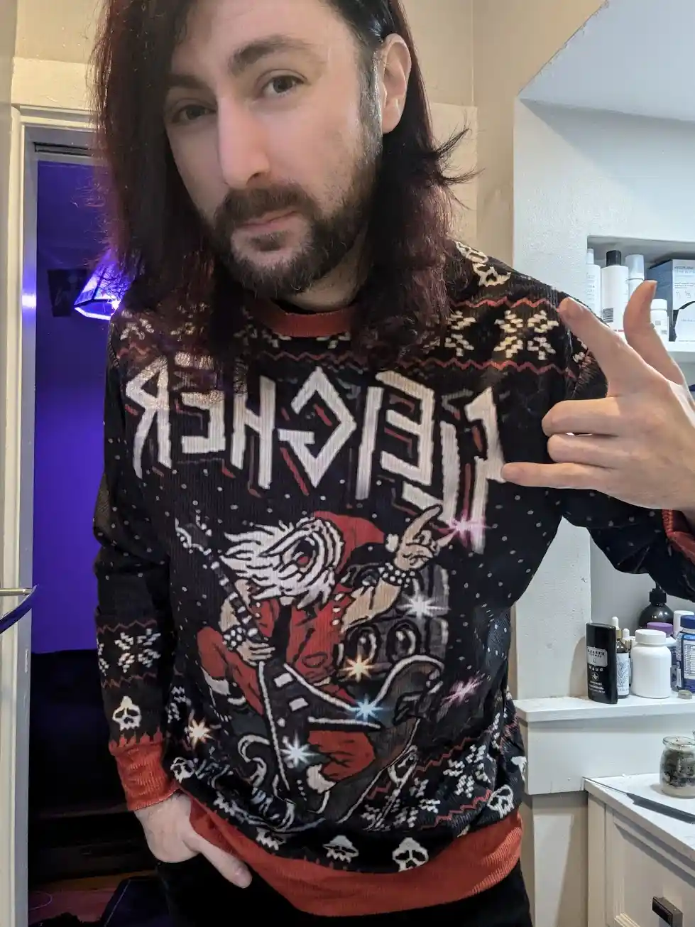 A man with long dark hair and a beard stands in a room, wearing a black holiday sweater with a graphic of Santa Claus…