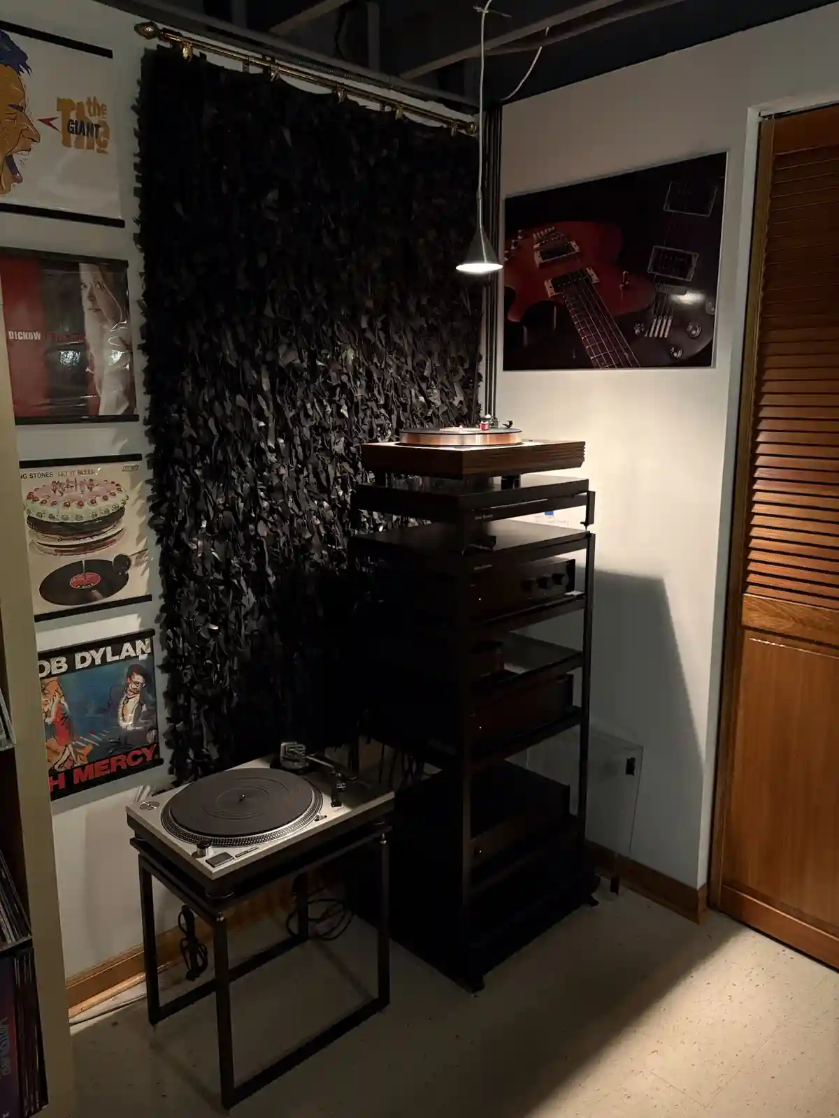 A dimly lit room features two record players: one on a small black metal stand and another on a tall black rack with audio…