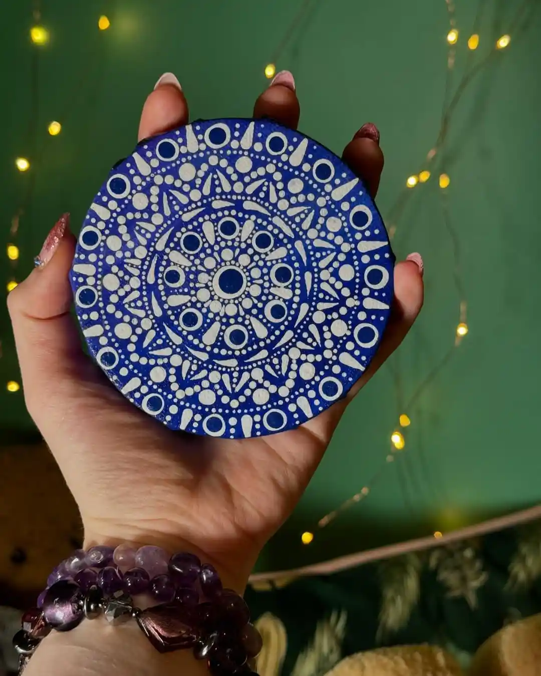A hand with decorated nails holds a circular blue and white mandala artwork against a green background with string lights…