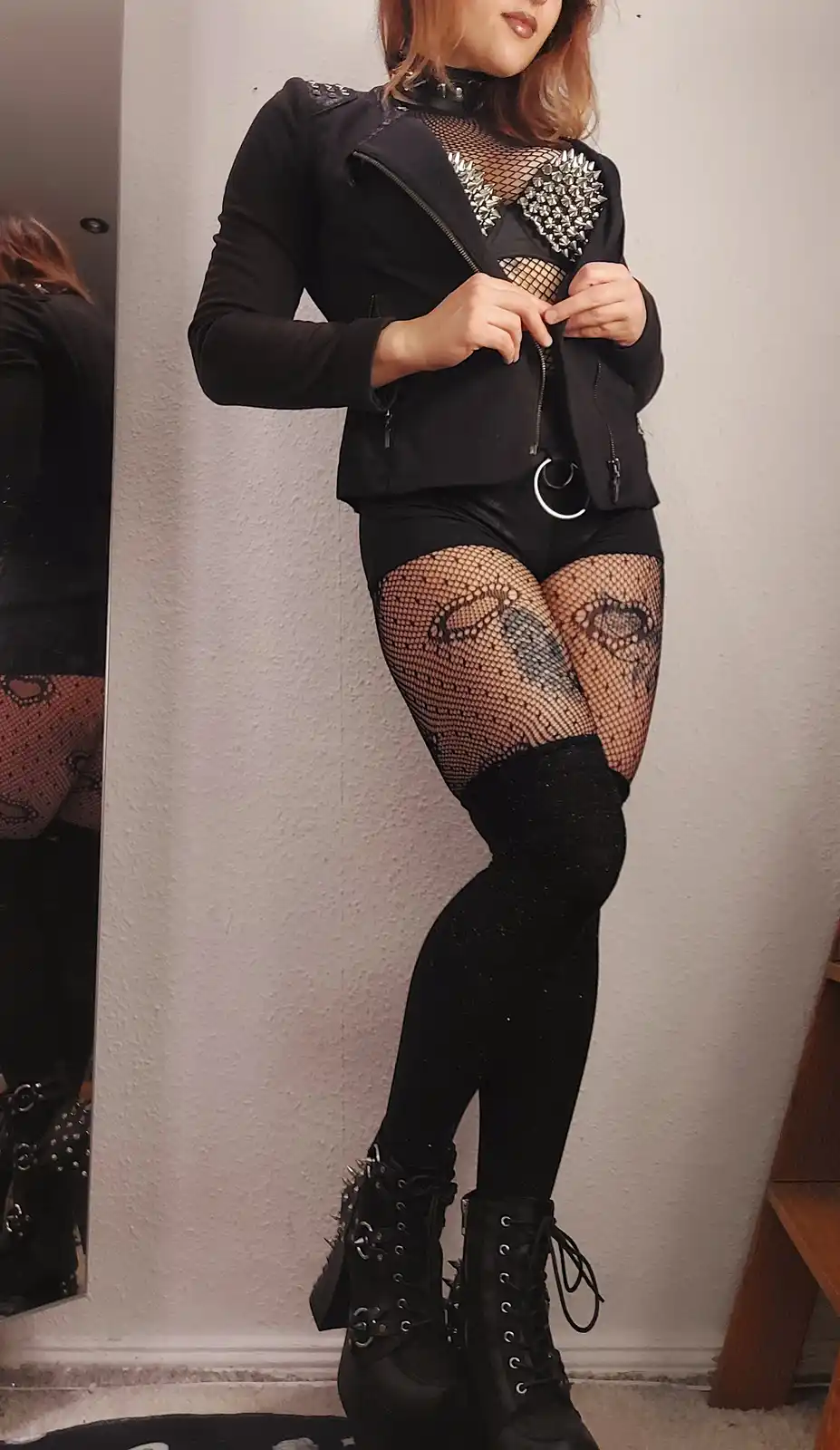 A person poses in a full-body mirror selfie wearing a black studded jacket, fishnet top, fishnet thigh-highs with skull…