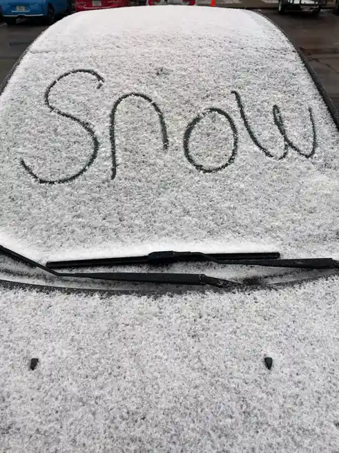 A car's windshield is covered in a layer of snow, with the word 'Snow' written in cursive in the snow using a dark marker or…