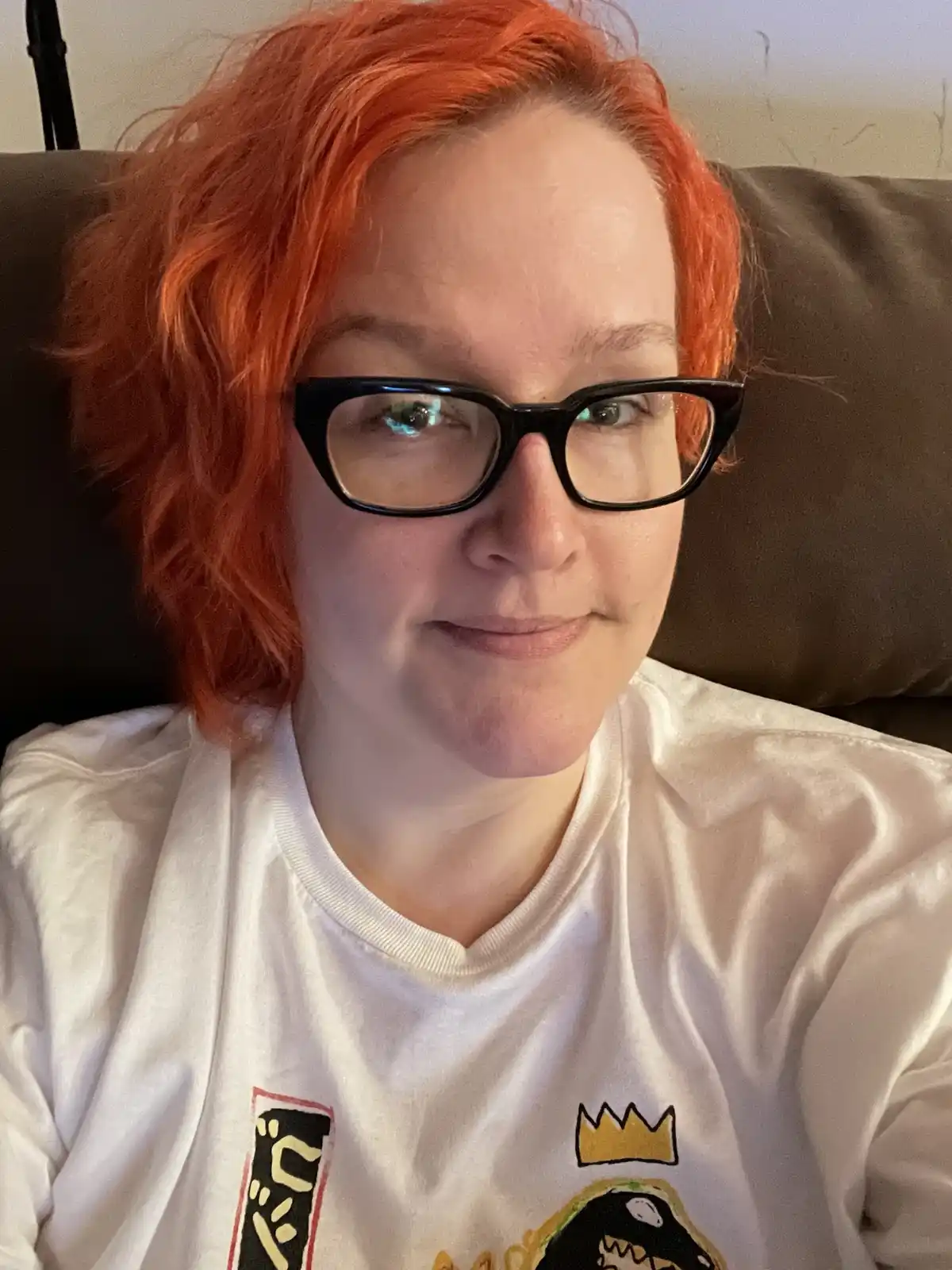 A person with short, vibrant orange hair and black-rimmed glasses sits on a brown couch, wearing a white t-shirt with a…