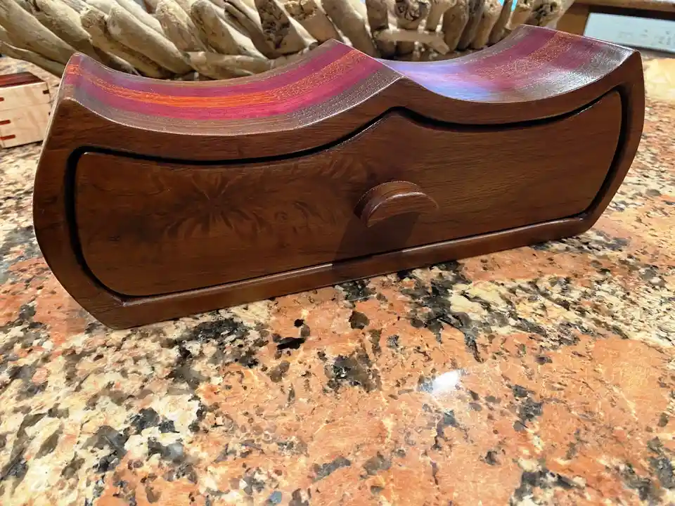 A wooden object with a curved top and a drawer, resembling a coffin, sits on a granite countertop. The top has a colorful…