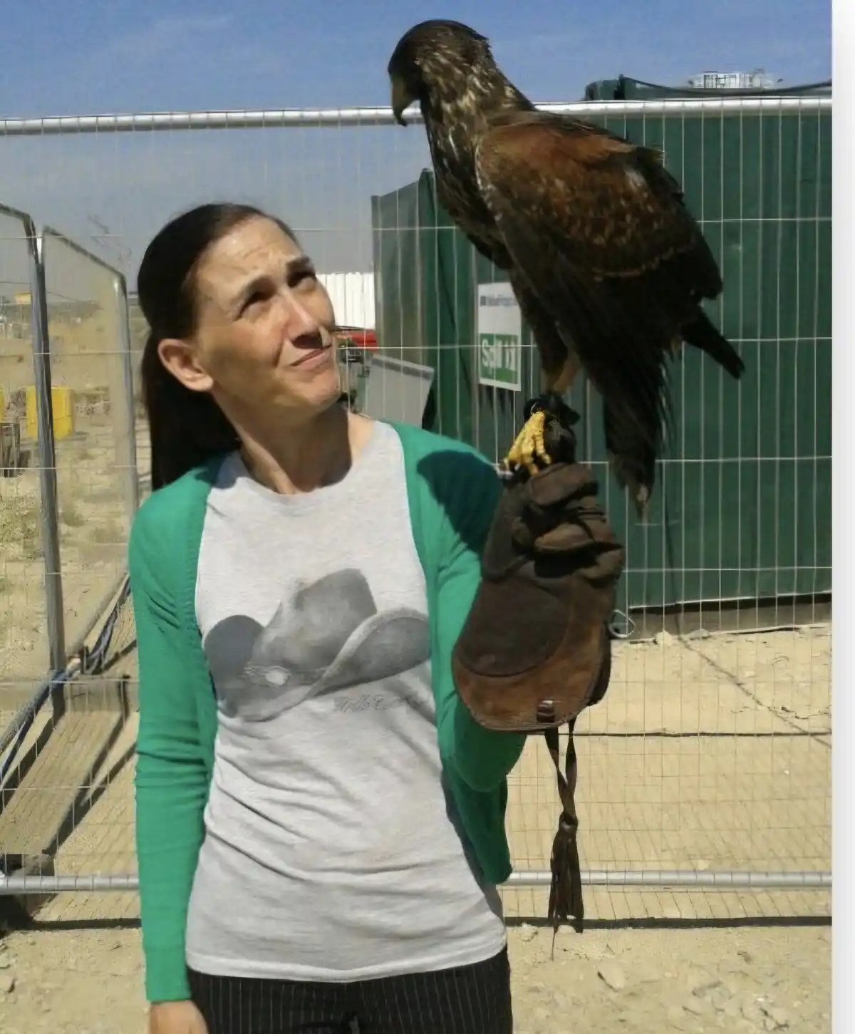 A woman wearing a green and grey shirt with a cowboy hat graphic stands outdoors, holding a large bird of prey, likely a…