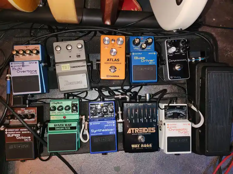 A dense, overhead shot of a musician's pedalboard filled with numerous guitar effects pedals from brands like Boss, Ibanez…