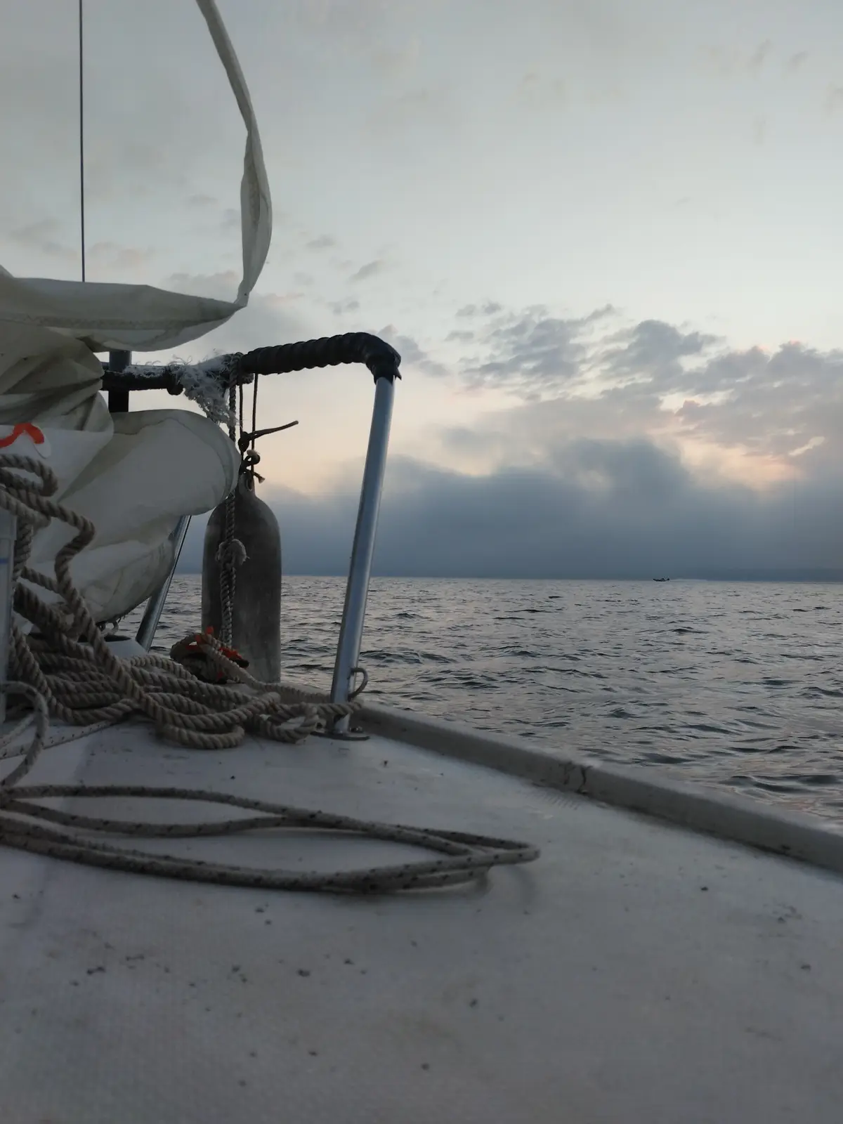 A serene view from the deck of a sailboat at dusk, showing coiled ropes, a metal rail, and a sail partially visible, with a…