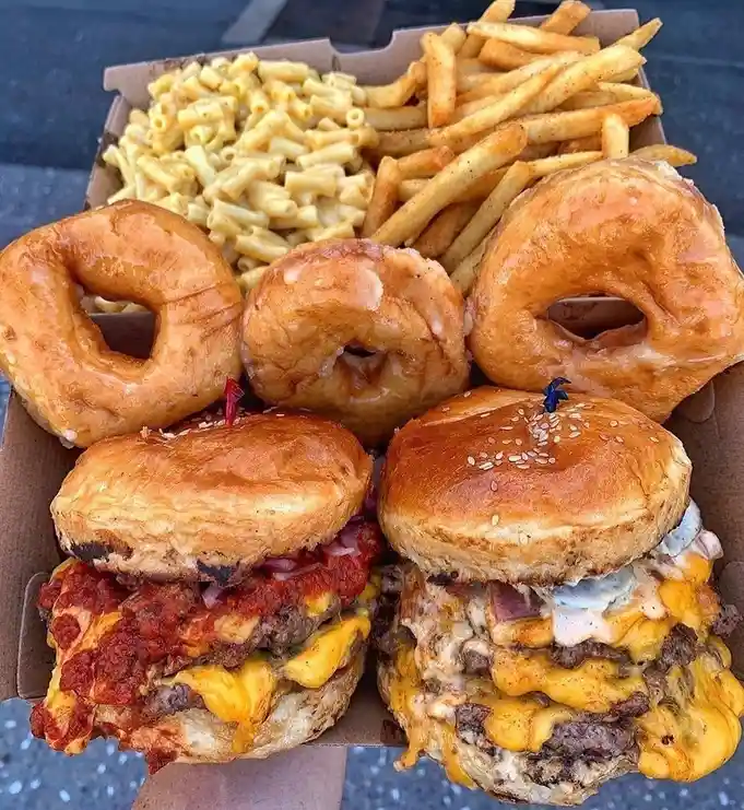 A cardboard food box contains two large cheeseburgers with toppings, three glazed donuts, french fries, and macaroni and…
