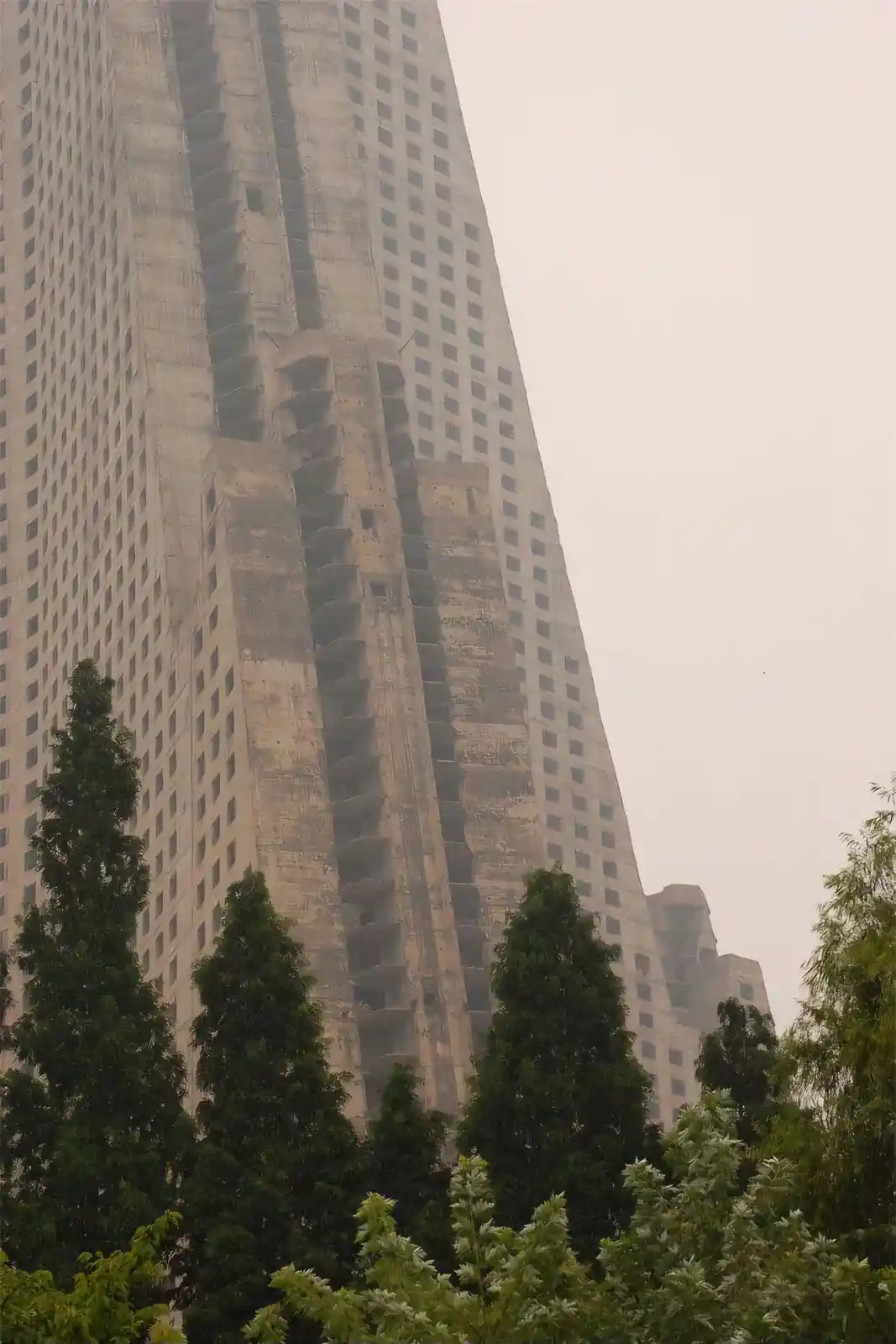 a tall, unfinished concrete skyscraper with visible structural damage and missing windows, viewed from below with green…