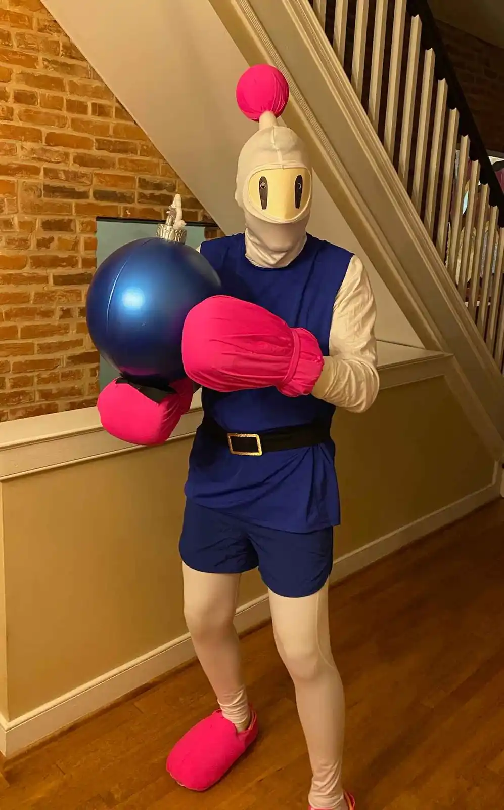 A person in a full-body costume resembling a video game character with a white mask, blue outfit, and pink boxing gloves, is…