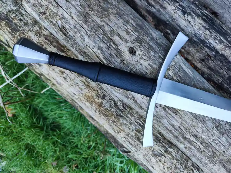 A silver-colored sword with a black wrapped grip and crossguard rests diagonally on a weathered wooden log, with green grass…