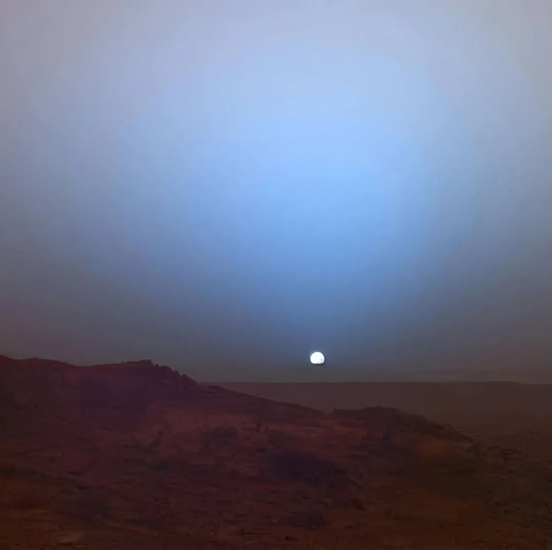 A wide shot of a barren, reddish-brown desert landscape under a vast sky, with a bright, full moon visible low on the…