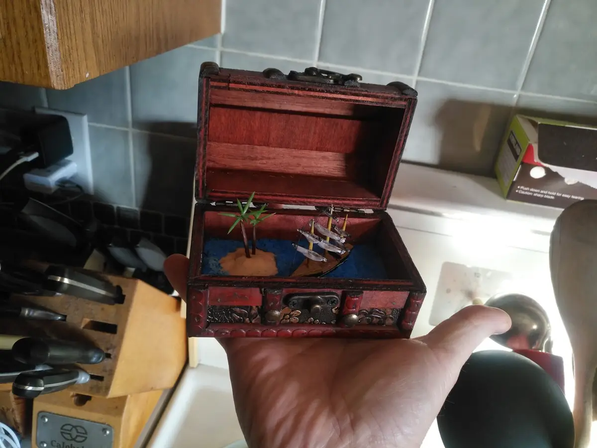 A hand holds a small, open wooden treasure chest with a miniature pirate ship and palm tree diorama inside, set against a…